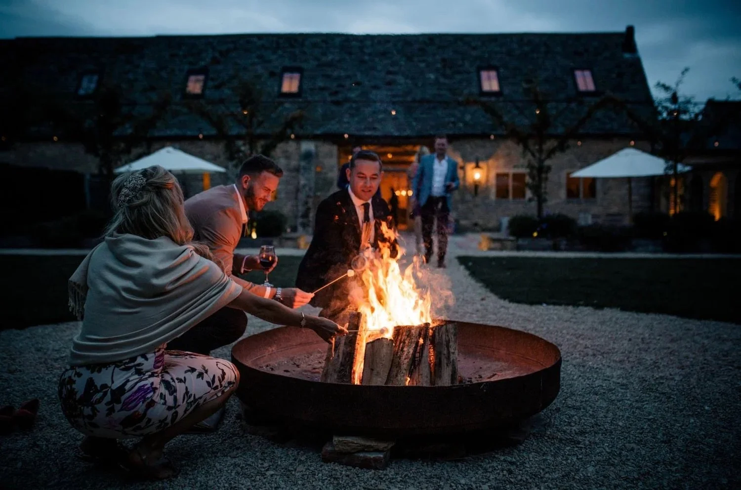 People gathered around a fire pit outside a house at dusk, some roasting marshmallows and holding wine glasses.