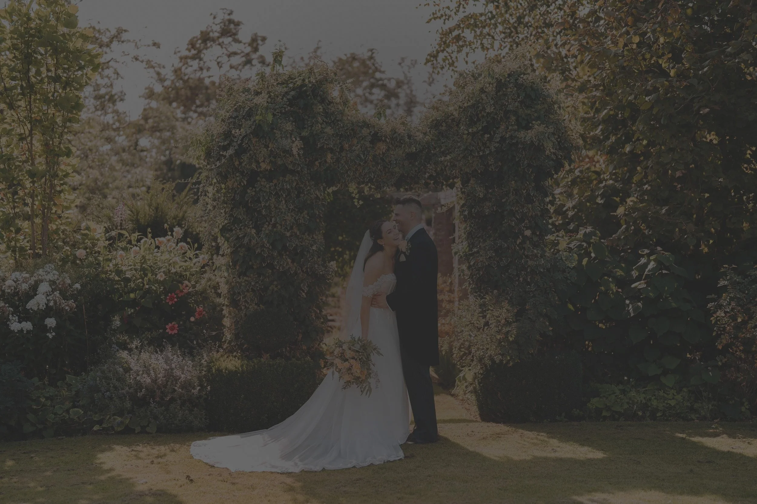 A bride and groom embrace under a floral arch in a garden.