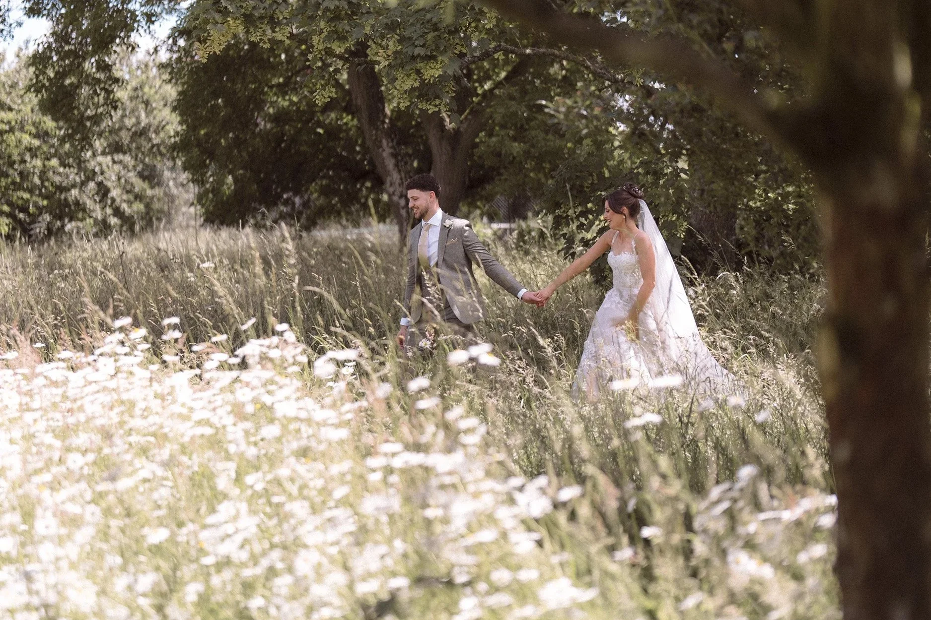 A bride and groom holding hands and walking through a grassy field filled with white wildflowers, with trees in the background on a sunny day.