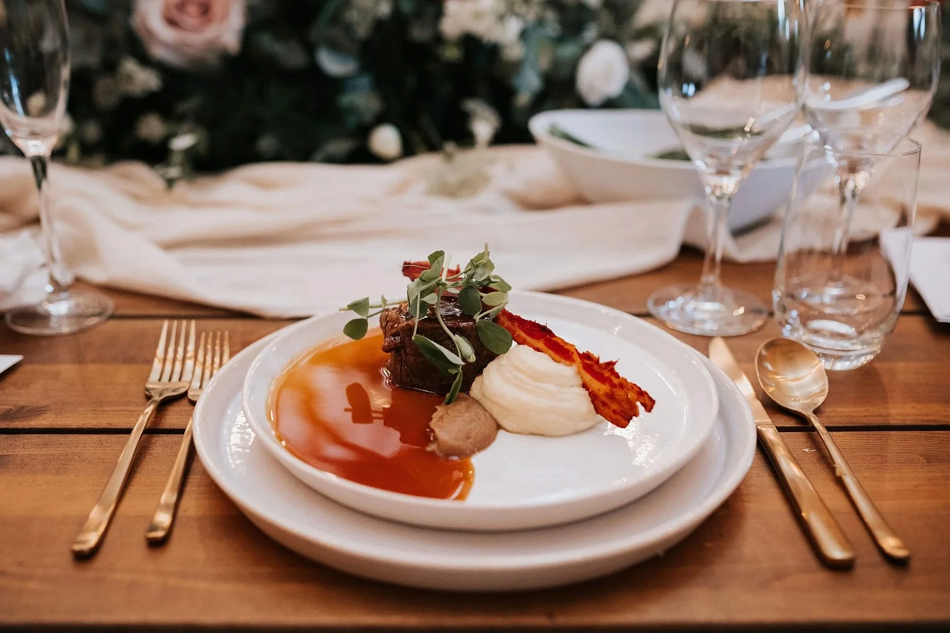 A plated gourmet meal on a white plate with mashed potatoes, a seared piece of meat garnished with herbs, crispy bacon strip, and sauce, set on a wooden table with gold utensils and wine glasses in the background.