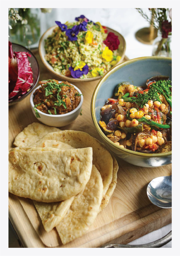 Plates of Indian food including naan bread, chickpea curry, vegetable stir-fry, and a bowl of rice garnished with edible flowers, on a wooden serving board.