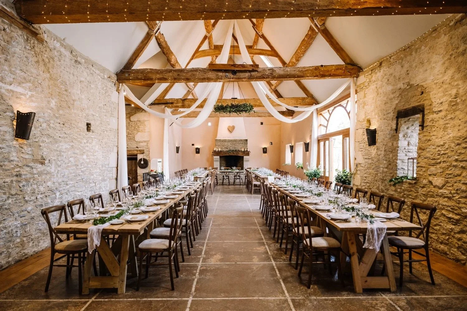 Beautiful rustic wedding reception setup with long wooden tables, chairs, and elegant table settings, inside a stone-walled venue with exposed wooden beams and large windows.