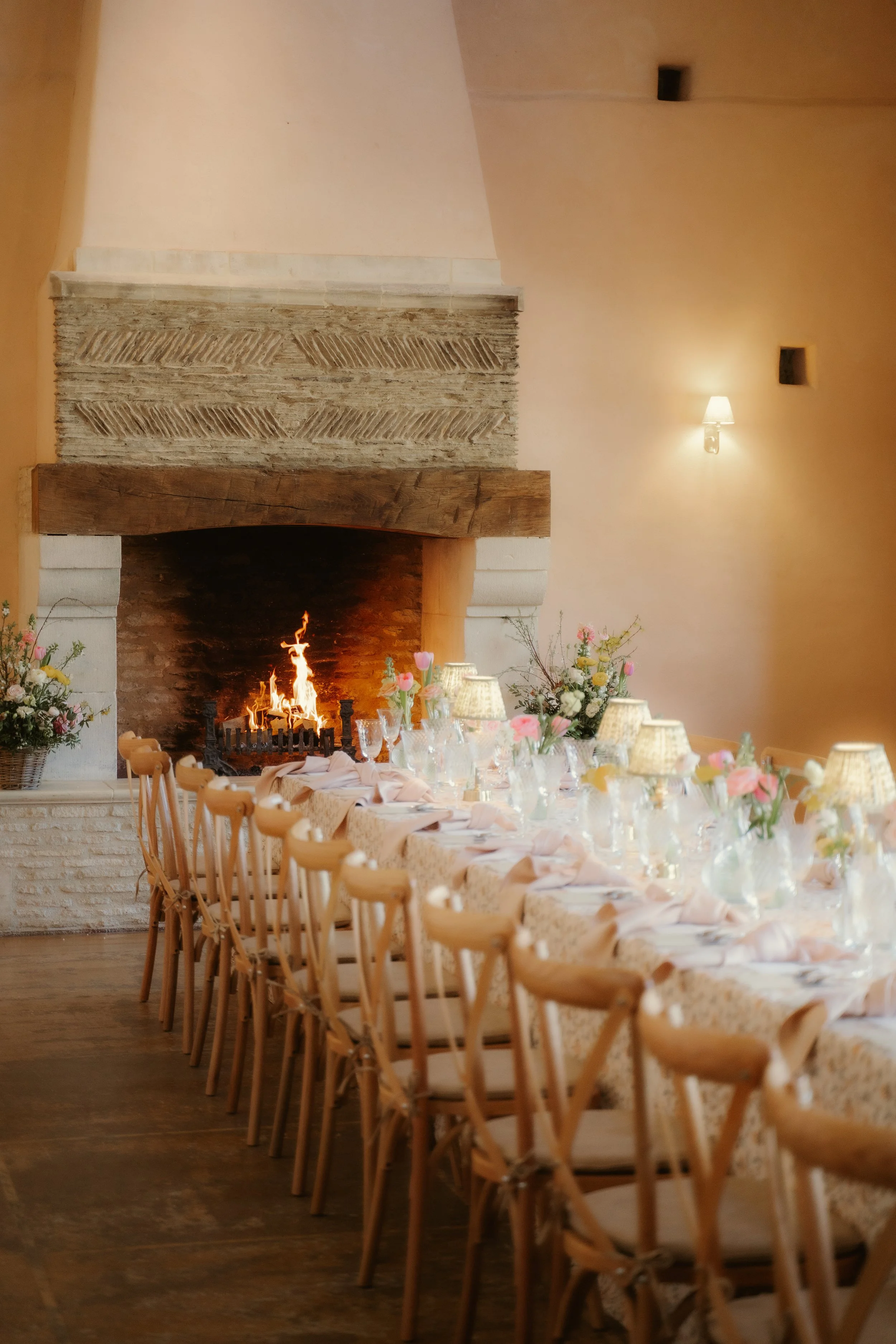 A long dining table set for a formal event, decorated with pink and yellow candles, floral arrangements, wine glasses, plates, and silverware, in a room with a brick fireplace in the background.