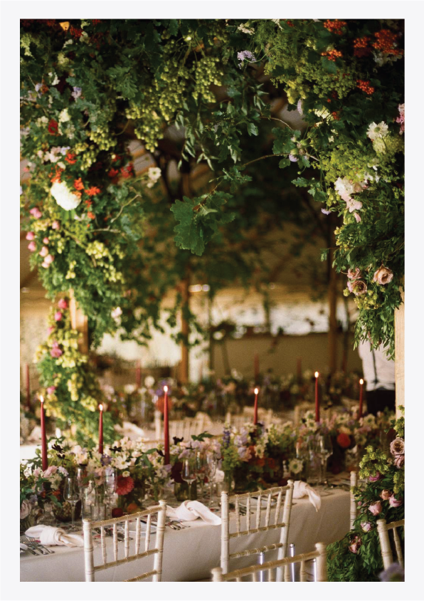 A decorated wedding or event table with floral arrangements, candles, and gold chairs underneath a lush floral arch.