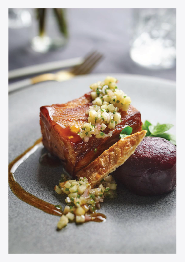 A plated gourmet dish featuring a piece of cooked pork belly topped with finely chopped apple and onion chutney, accompanied by roasted beet and green garnish, artistically presented on a gray plate.