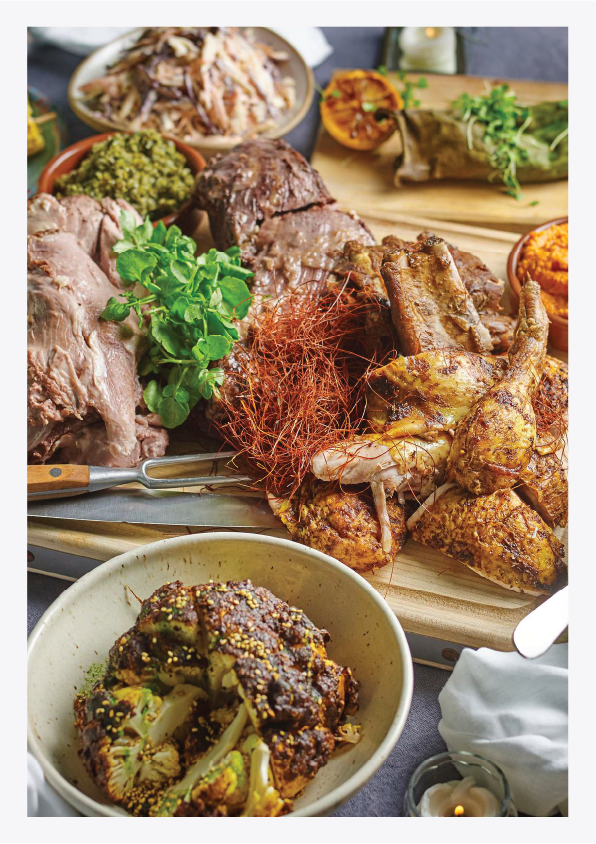 A spread of various foods including roasted meats, greens, roasted vegetables, and side dishes on a table. There is a cutting board with roasted chicken, a bowl of roasted cauliflower, a plate with sliced beef, and containers with greens, mashed pota