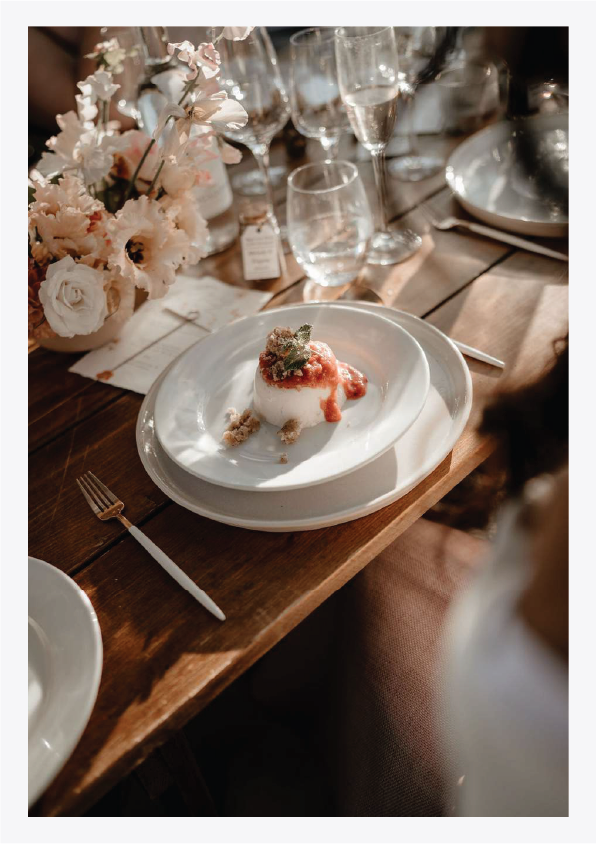 A plated cheesecake dessert topped with fruit and mint, surrounded by glasses of water and sparkling wine, on a wooden dining table with a pink flower centerpiece.