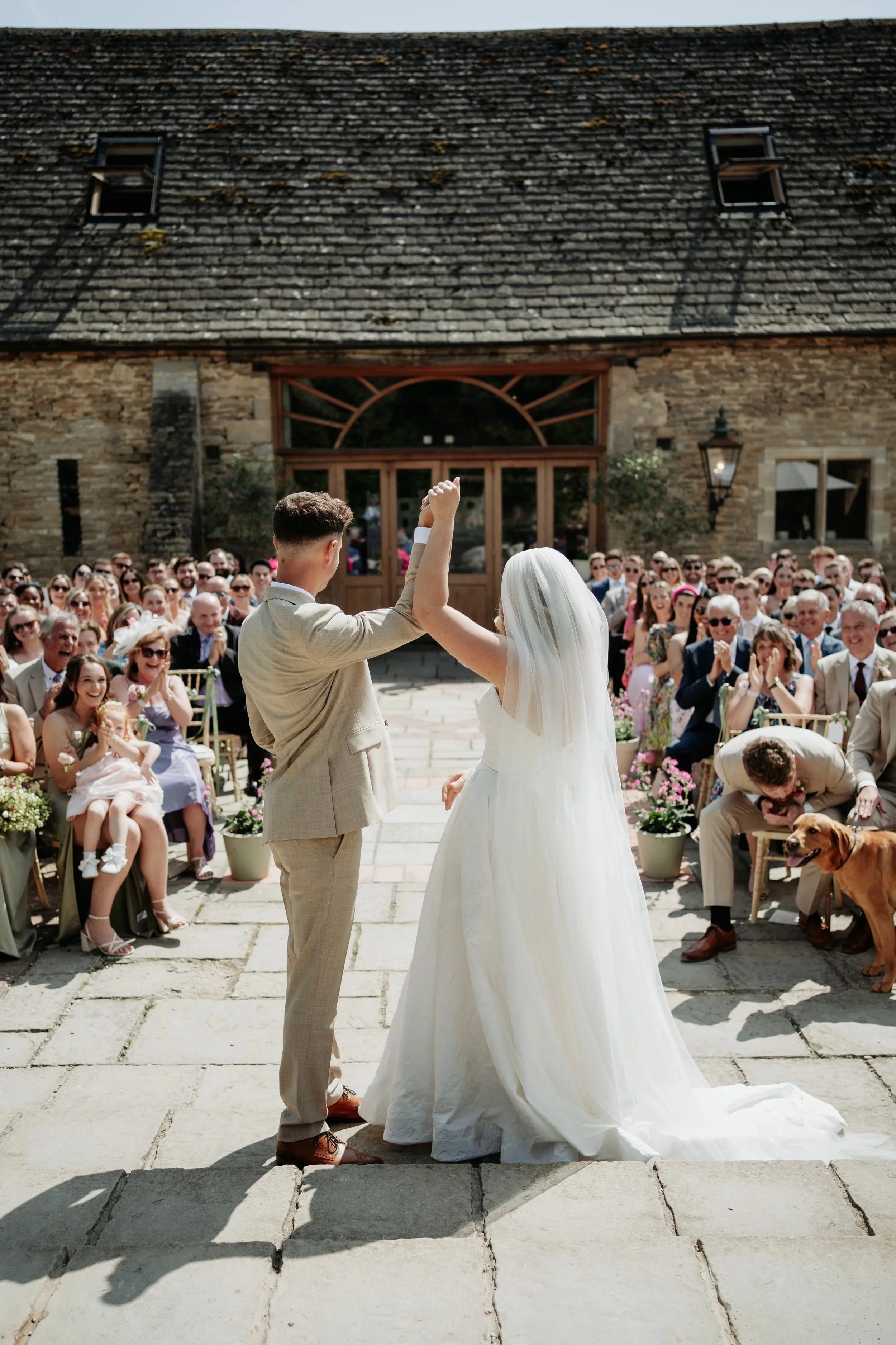 A couple is getting married outdoors, with many guests seated on both sides, some clapping and smiling, in front of a rustic brick building with a large glass door. The bride and groom are holding hands and appear happy.