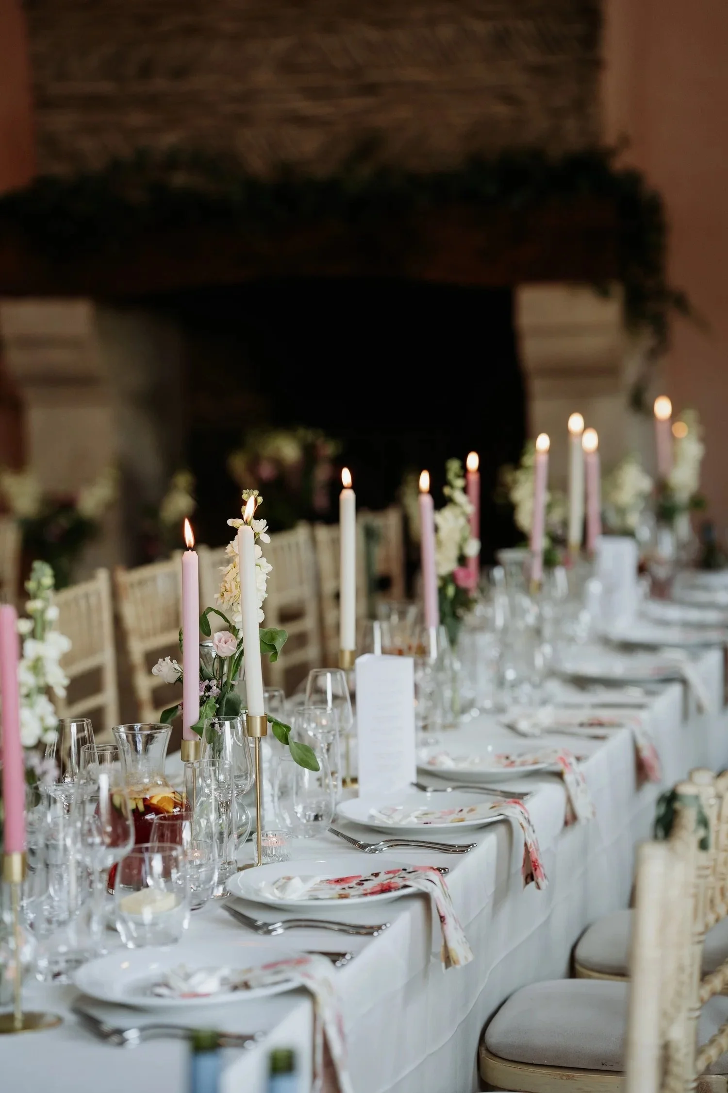 A long dining table set for a formal event, decorated with pink and white candles, floral arrangements, wine glasses, plates, and silverware, in a room with a brick fireplace in the background.