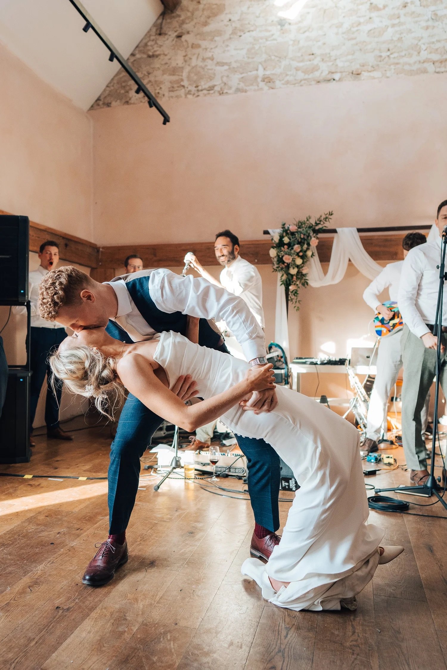 A wedding reception scene with a bride and groom dancing. The groom is dipping the bride backwards in a dance pose while people in the background celebrate and play music.