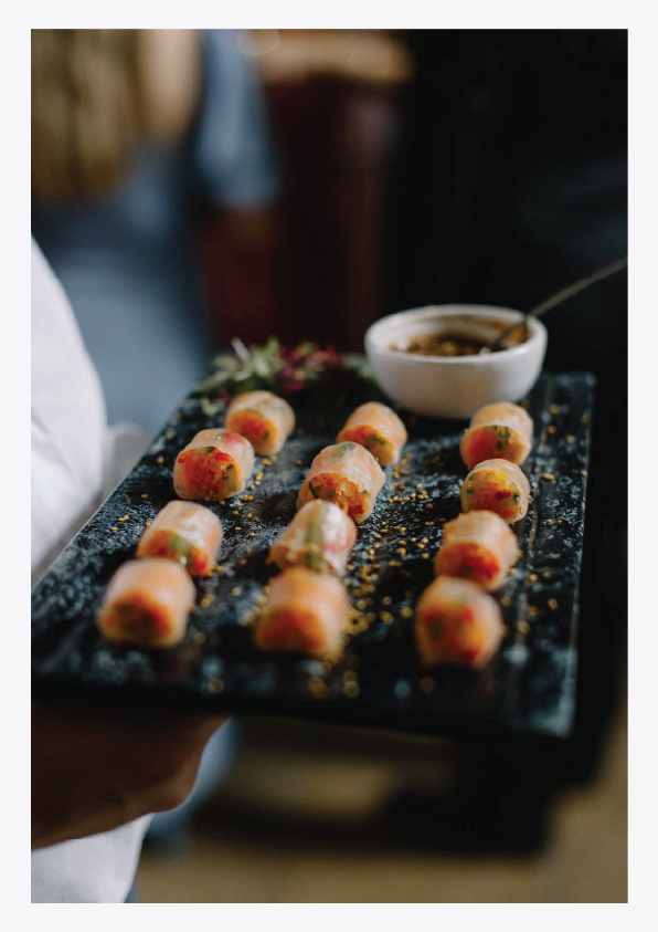 A person holding a black rectangular platter with small, colorful, uncooked sushi rolls and a white bowl of dark dipping sauce.