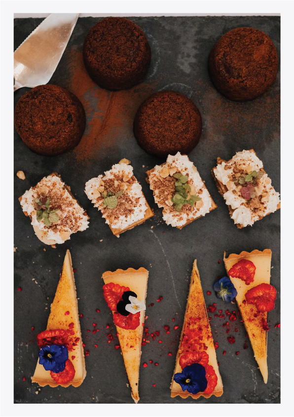 Assorted desserts on a dark serving tray, including four chocolate truffles, four square layered cakes topped with crumble and mint, and four triangular cheesecakes decorated with berries, edible flowers, and red sprinkles.