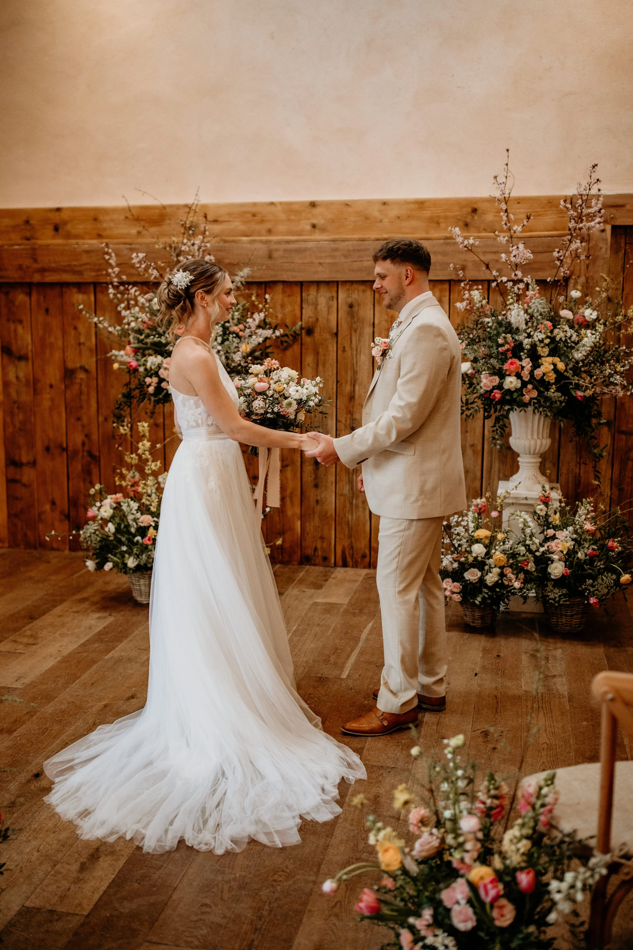 A couple is getting married inside, with many guests seated on both sides, some clapping and smiling, inside a rustic barn with a large glass door. The bride and groom are holding hands and appear happy.