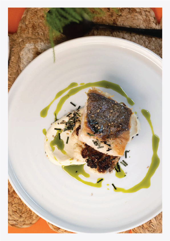 A plated dish of cooked fish fillet with crispy skin, served with mashed potatoes, garnished with chopped herbs and drizzled with green sauce.