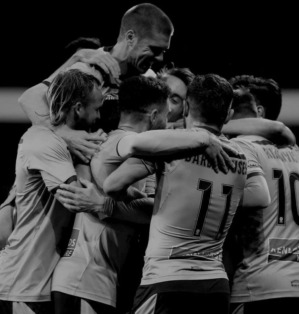 A group of soccer players huddled together in a celebratory hug on the field with a coach also joining in the embrace.