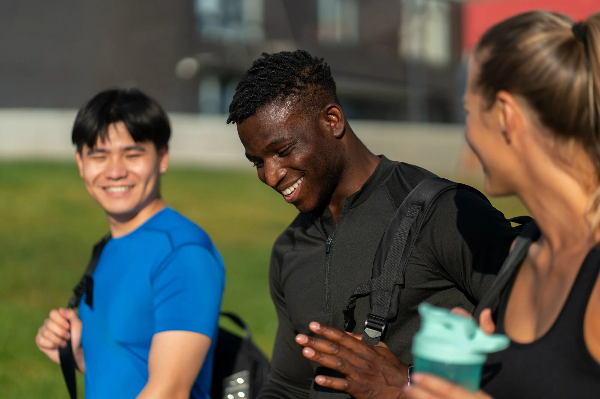 Three people, two men and one woman, standing outside having a conversation. They are dressed in athletic clothing and carrying backpacks. The woman is holding a teal water bottle. They are smiling and looking at each other.