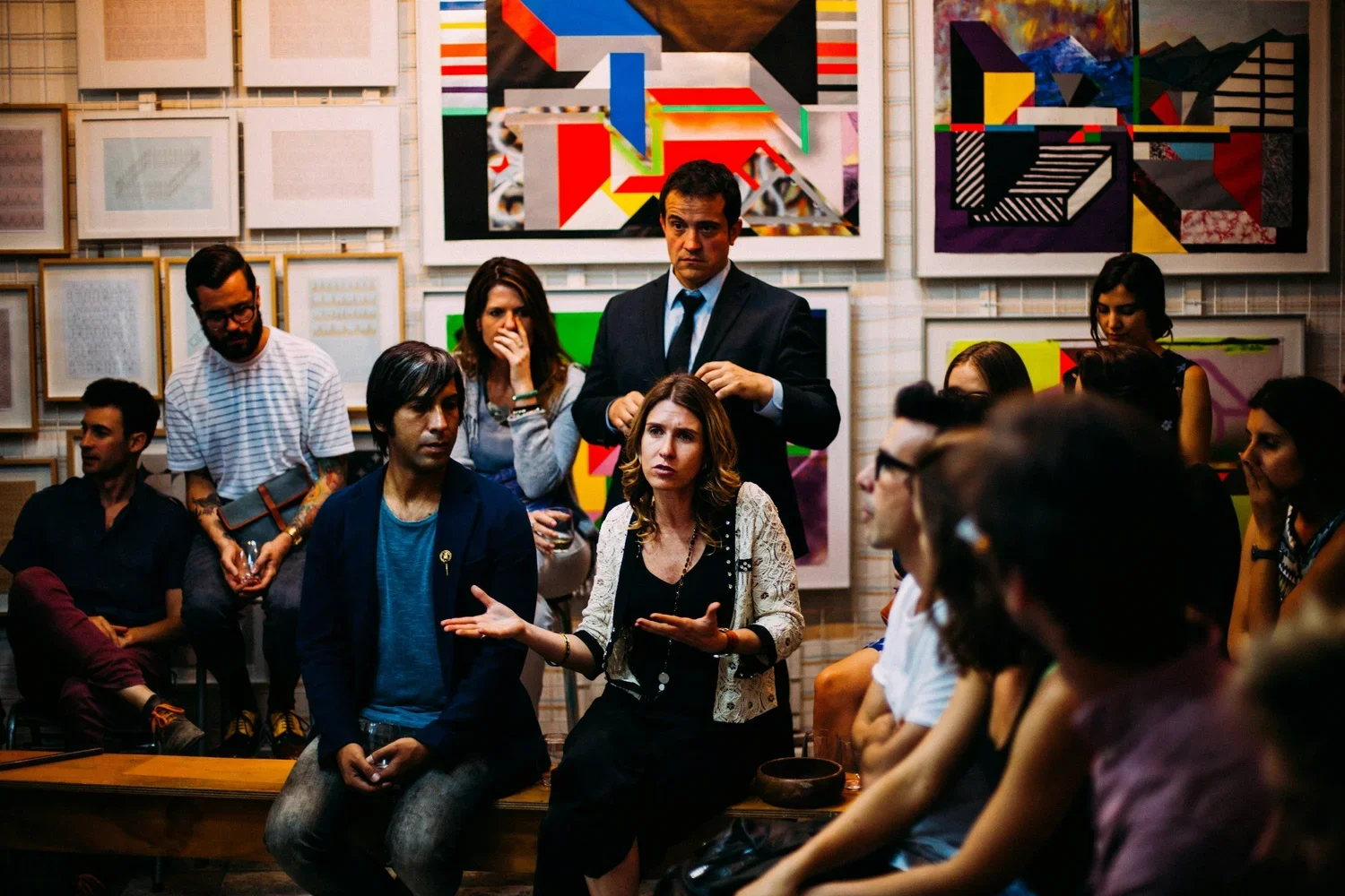 A group of people in an art gallery listening to a woman speak. The background features colorful abstract artwork on the walls.