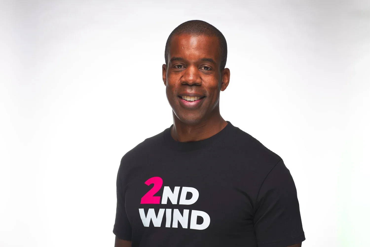 A smiling man wearing a black T-shirt with '2ND WIND' written on it in bold letters, against a plain white background.