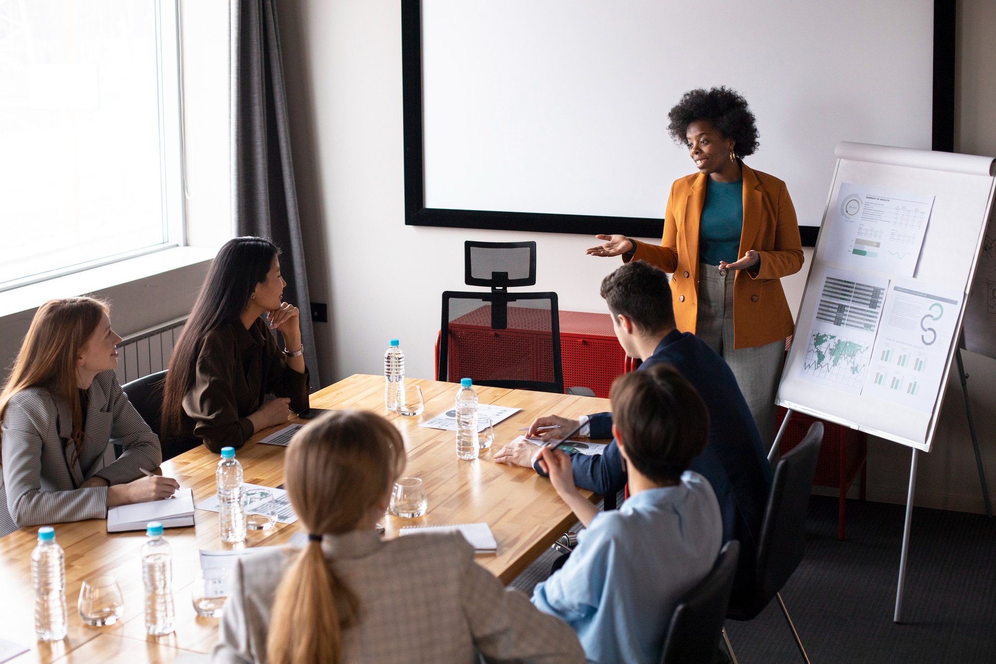 A woman giving a presentation to a group of five colleagues in a conference room, with charts on a whiteboard and a large screen behind her.