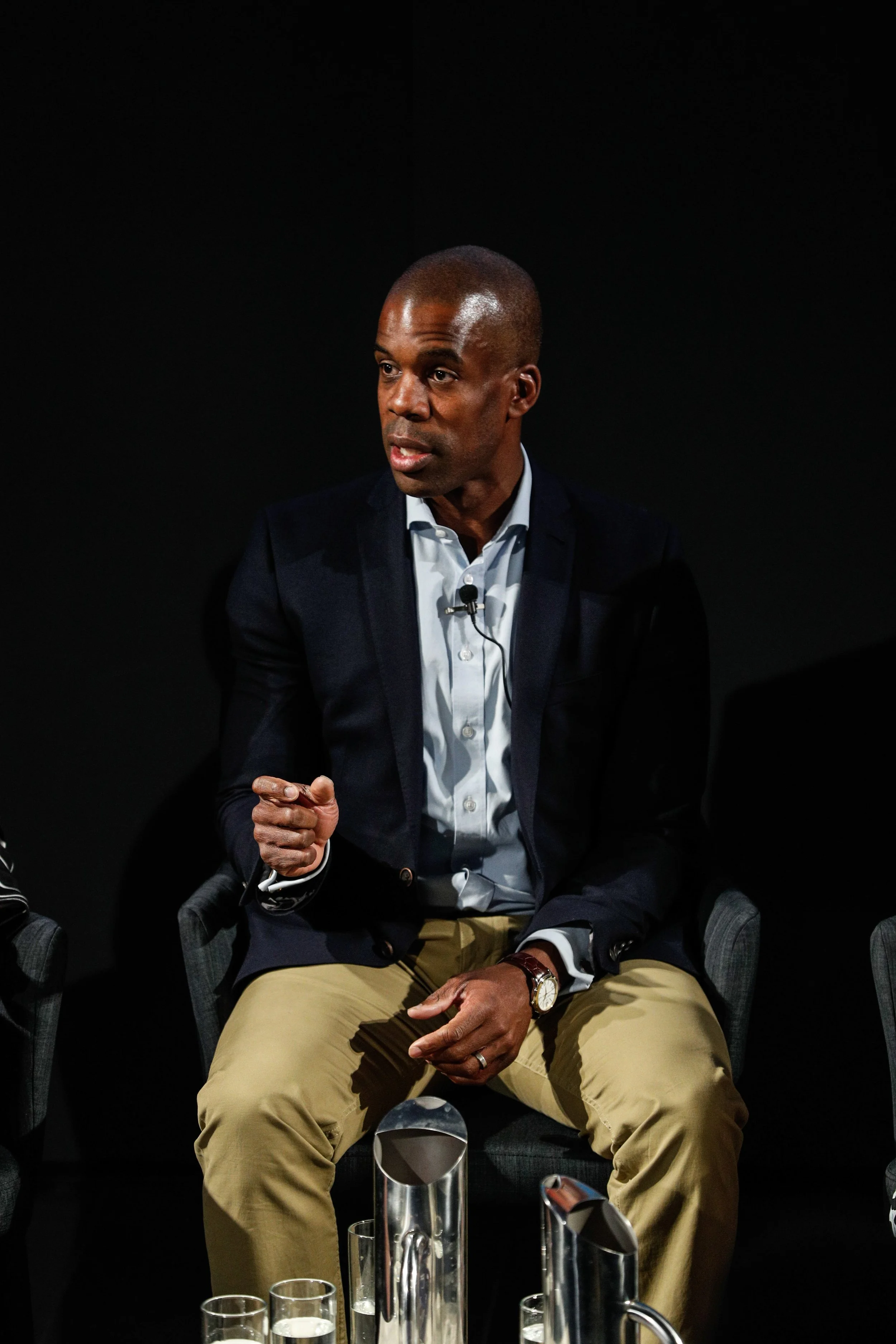 A man wearing a dark blazer and khaki pants sitting on a chair, speaking into a microphone, in front of a black background.