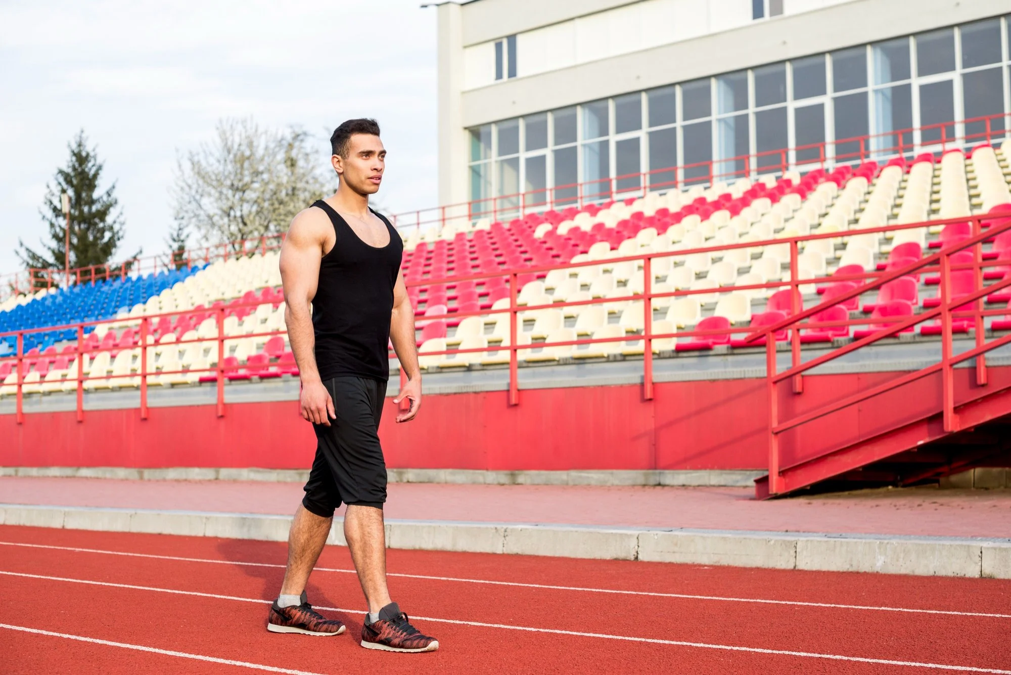 Young male athlete walking on a red running track at an outdoor stadium with empty red, white, and blue spectator seats.