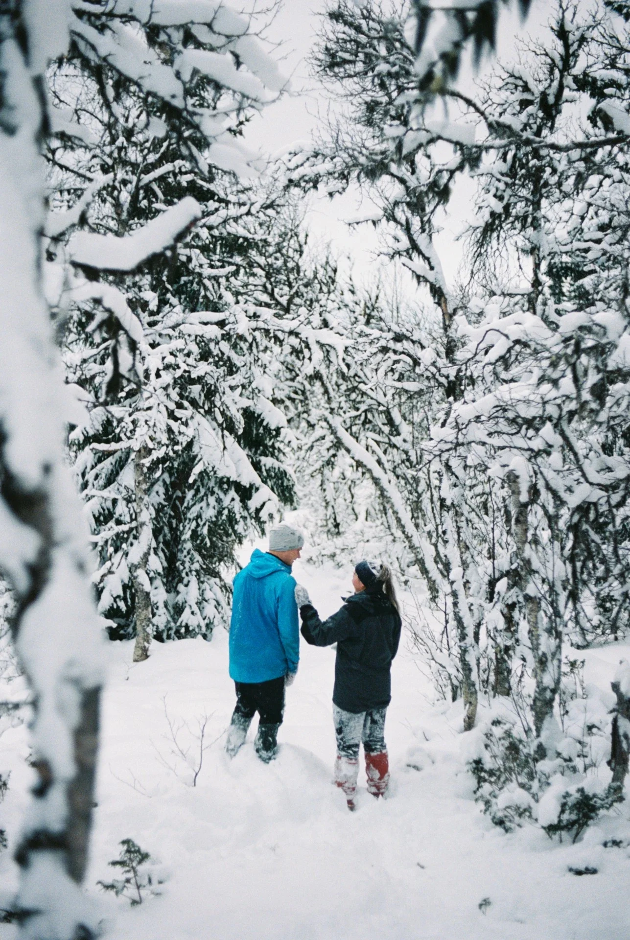 To personer går gjennom en snødekt skog, kledd i vinterklær, med trær dekket av snø rundt seg.