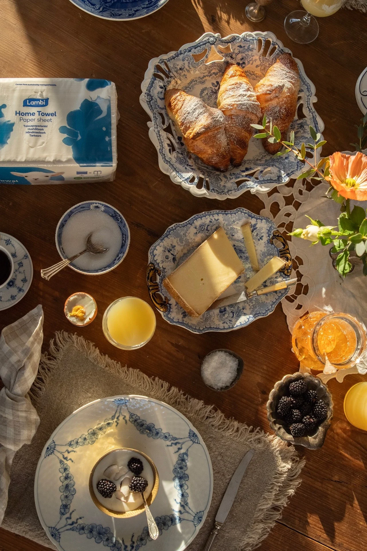 En dansk morgenbuffé med croissanter, ost, pisket krem, blackberries, juice, og kjeks på en tregulv med serviett og blomster.