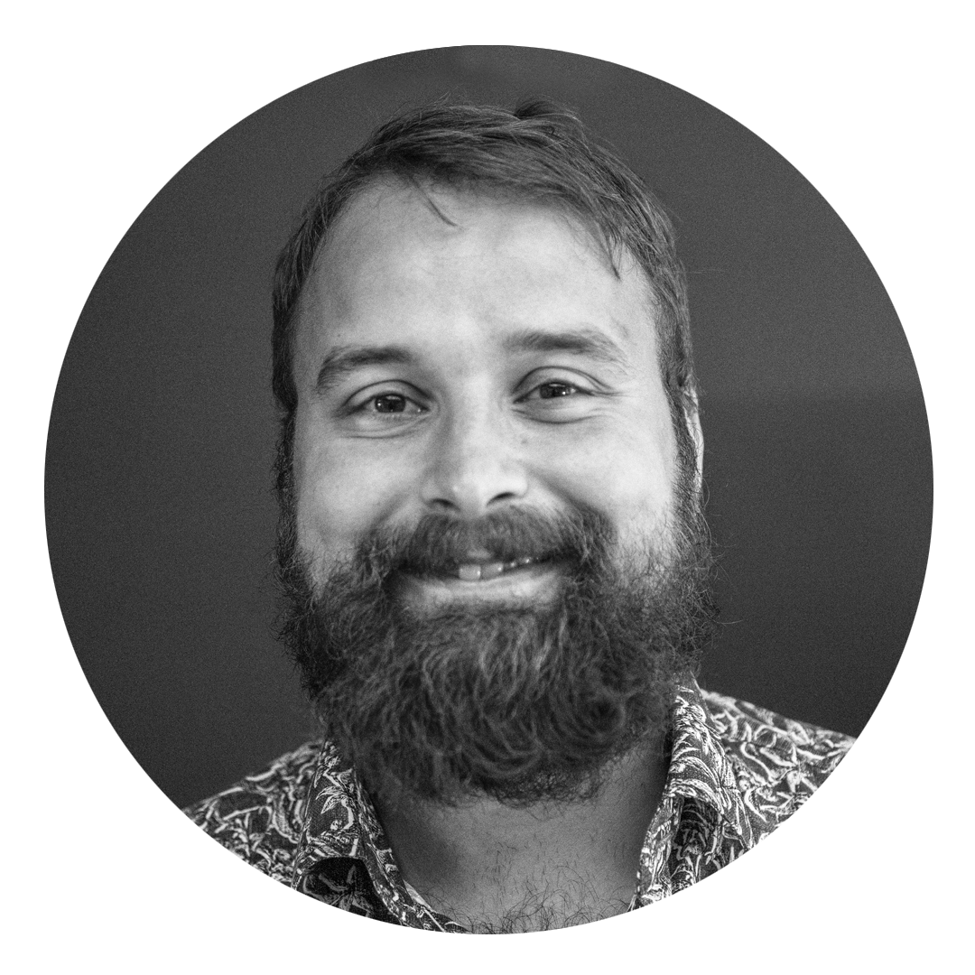 Svart-hvitt portrett av en mann med langt skjegg og kort hår, smilende, iført en patterned skjorte.