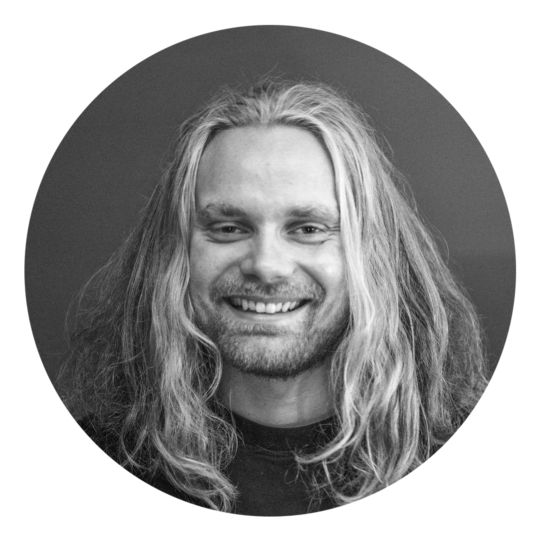 Svart-hvitt portrett av en smilende mann med langt, blond hår og skjegg, mot en enkel bakgrunn.
