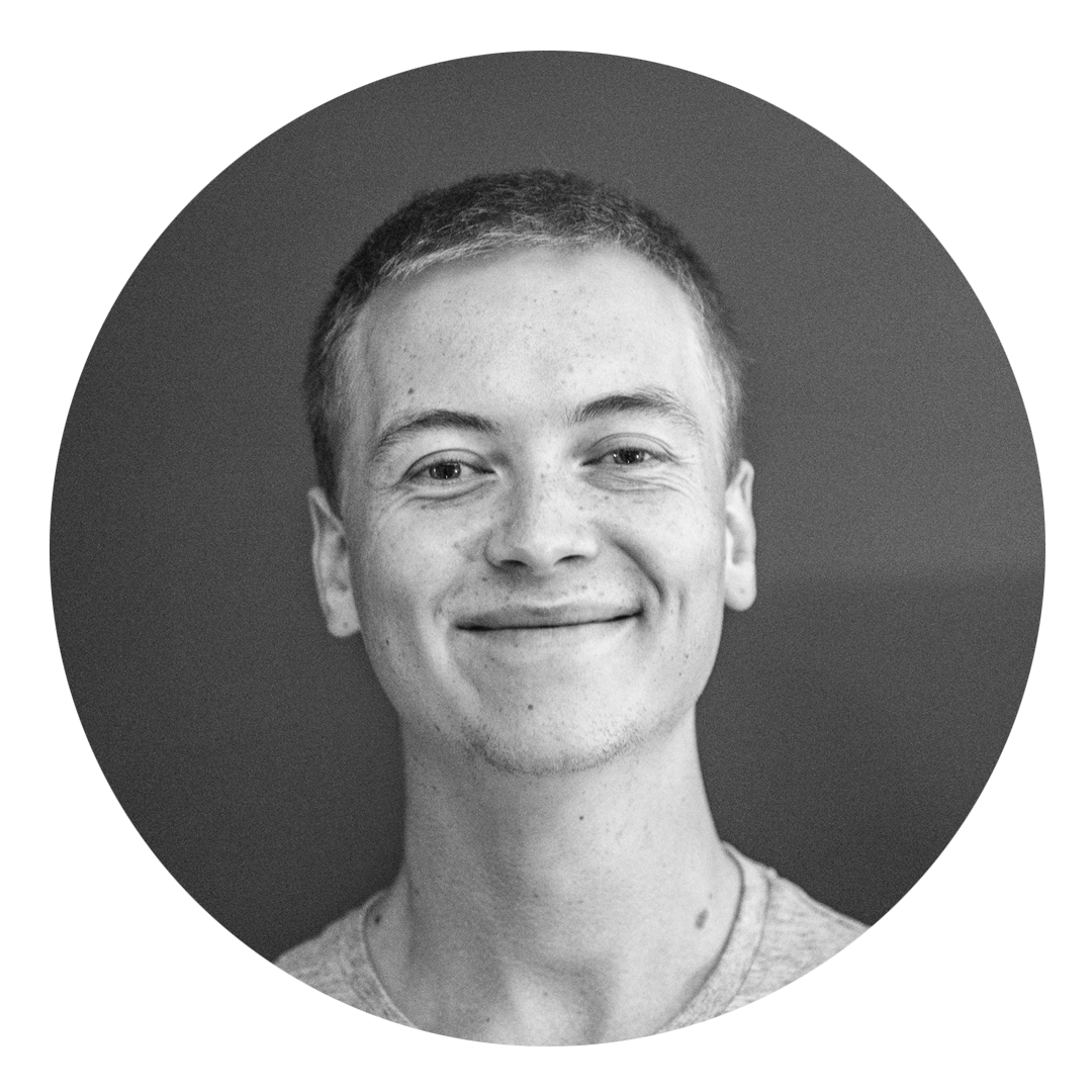 Svart-hvitt portrett av en smilende ung mann med kort hår og lett fregnet hud, mot en mørk bakgrunn.