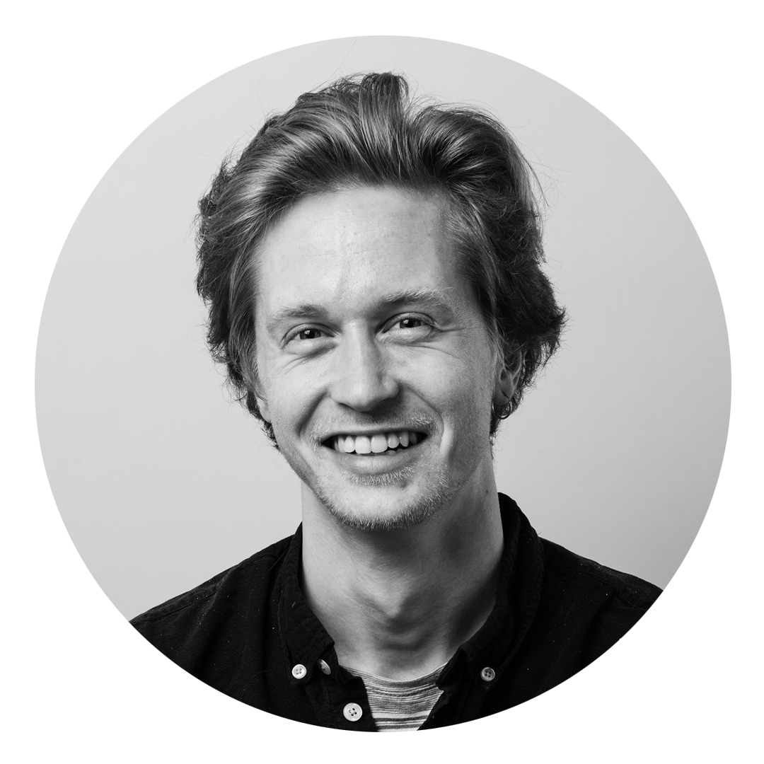 Sort-hvitt portrett av en smilende ung mann med lyst hår, iført svart skjorte, fotografert i studio.