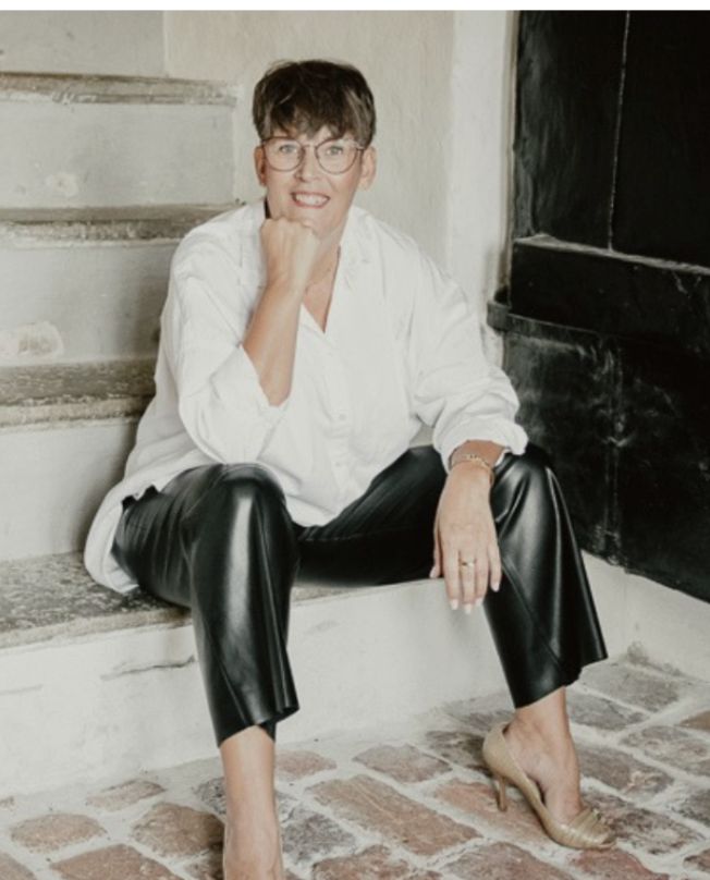 Kvinna i vit blus och svarta läderbyxor sittande på en trappa, ler.