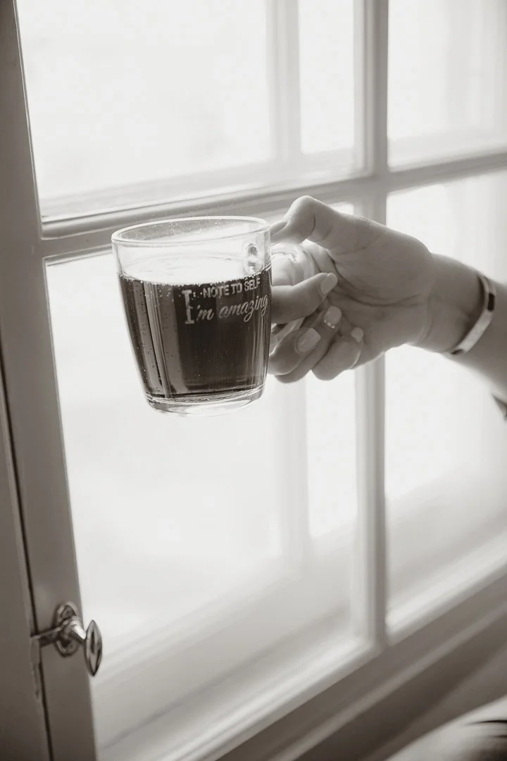 En hand håller i ett glas med mörk dryck framför ett fönster.