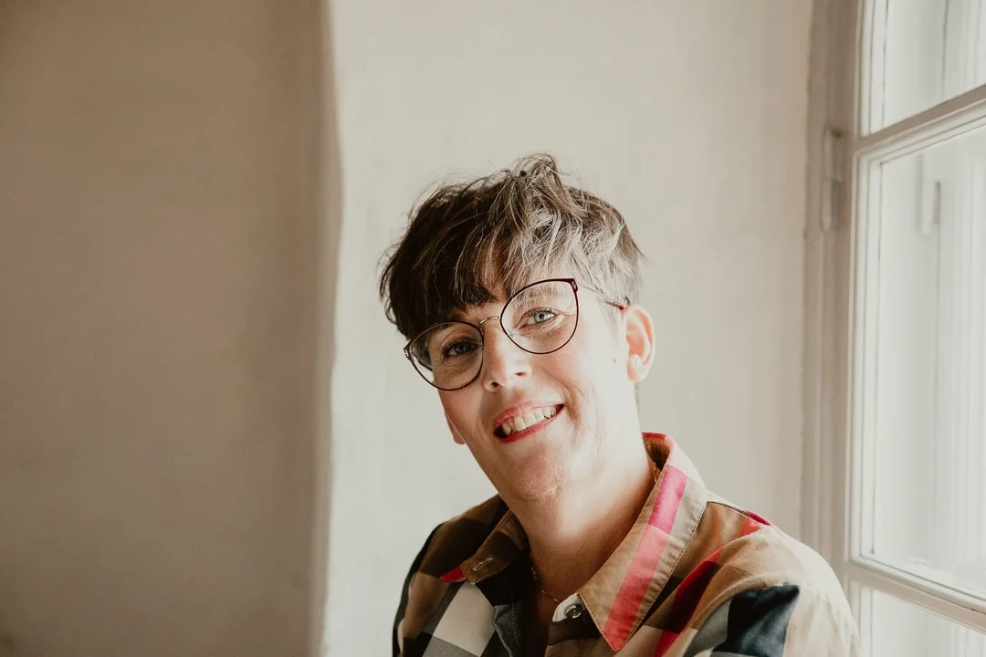 En person med kort, vågigt hår och glasögon, ler mot kameran, står bredvid ett fönster.