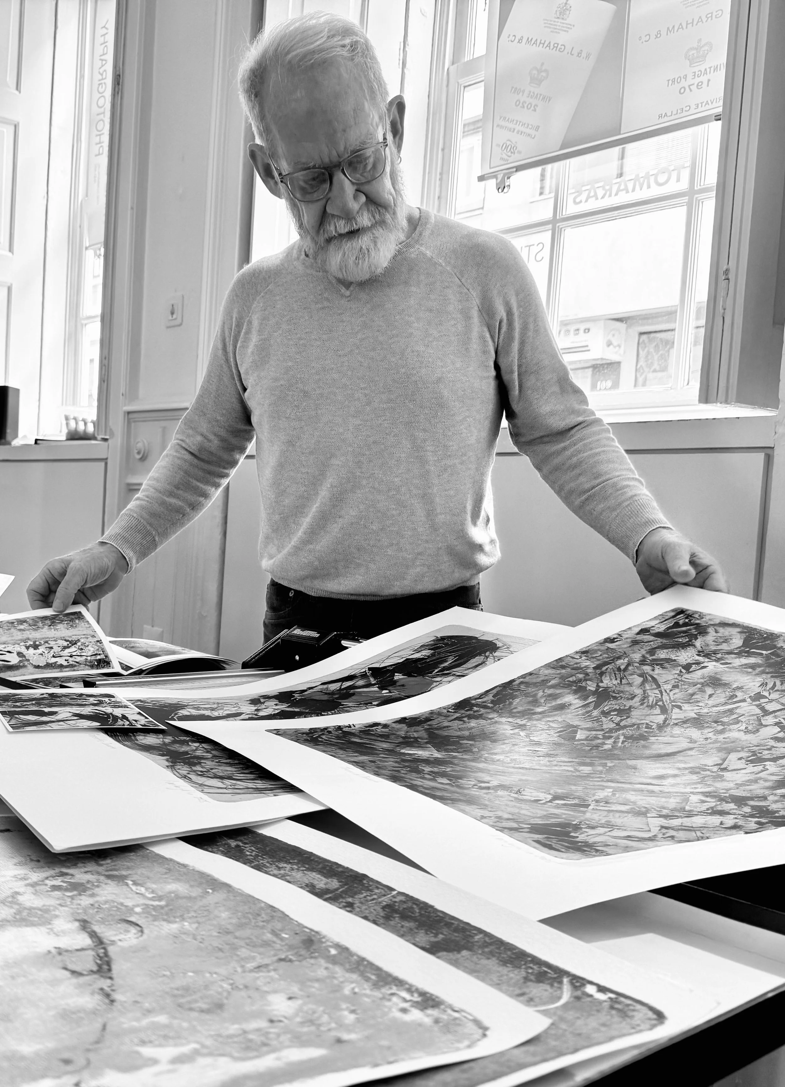An elderly man with glasses and a beard is looking at and sorting through large printed photographs spread out on a table inside a well-lit room with large windows.