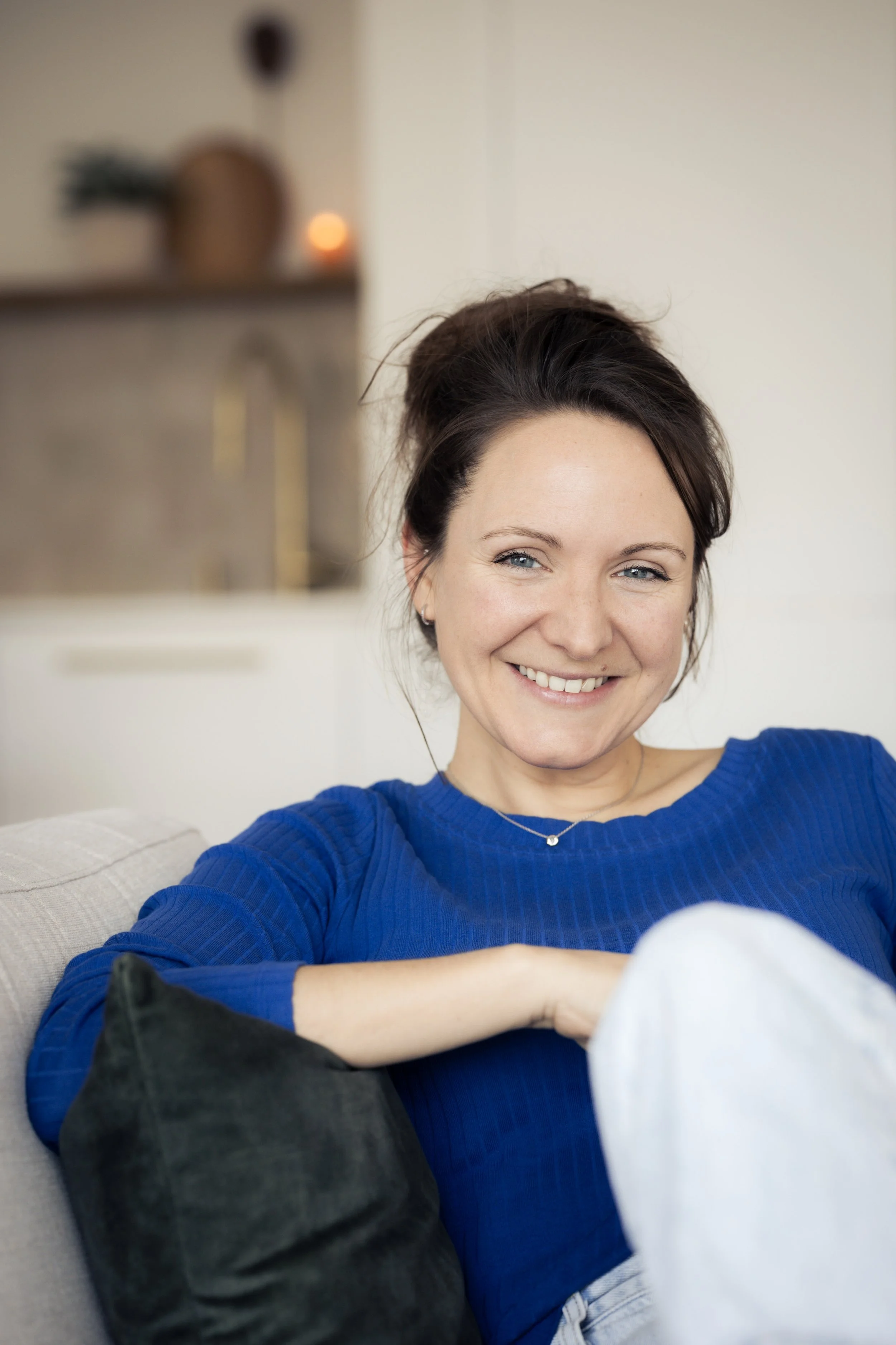 Une femme souriante assise sur un canapé, portant un vêtement bleu et entourée de coussins, dans un intérieur chaleureux.