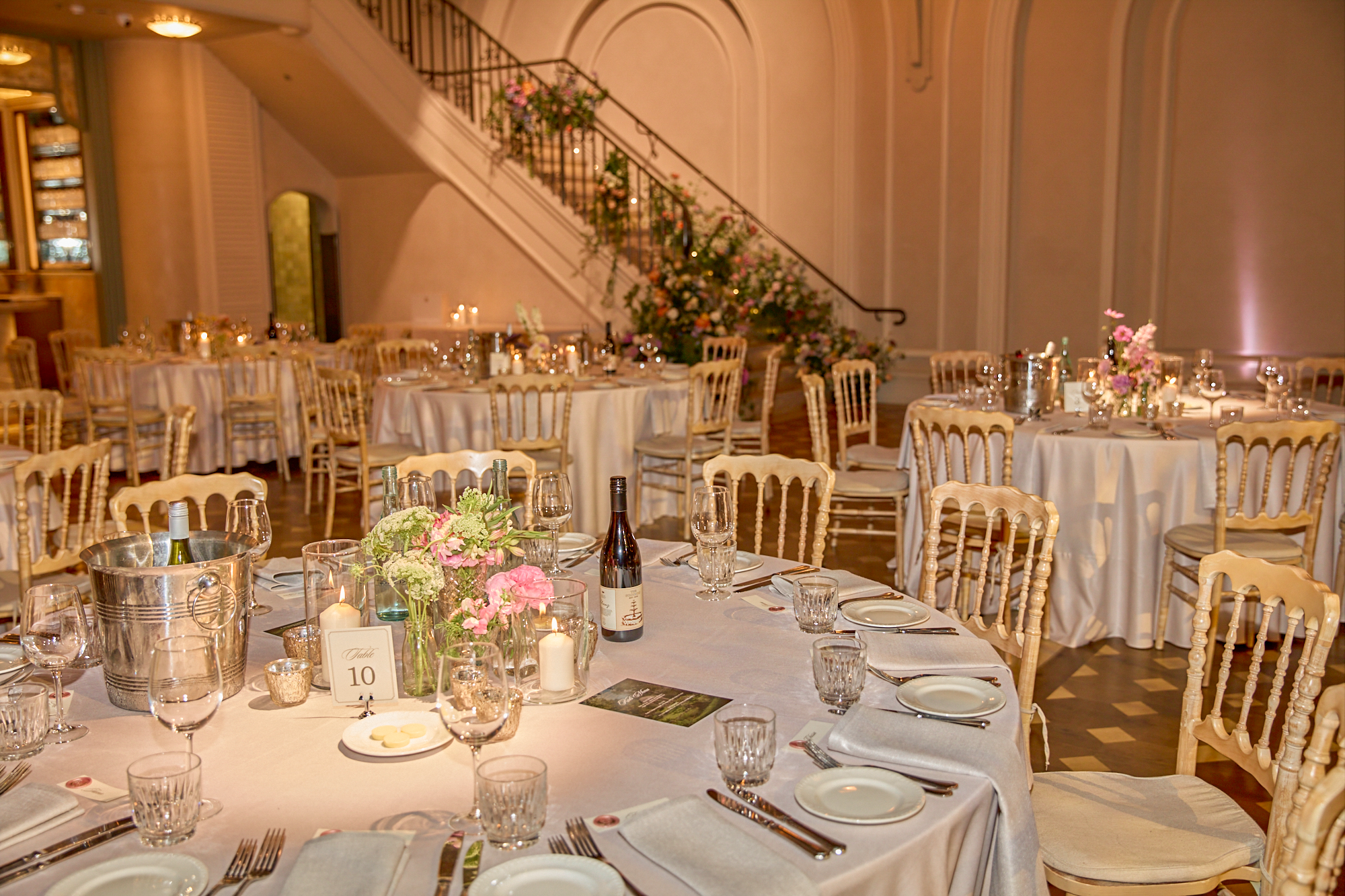 Elegant banquet hall set up for an event with round tables covered with white tablecloths, decorated with floral centerpieces, candles, wine, and glassware; wooden chairs surround the tables; a staircase with floral arrangements is visible in the bac