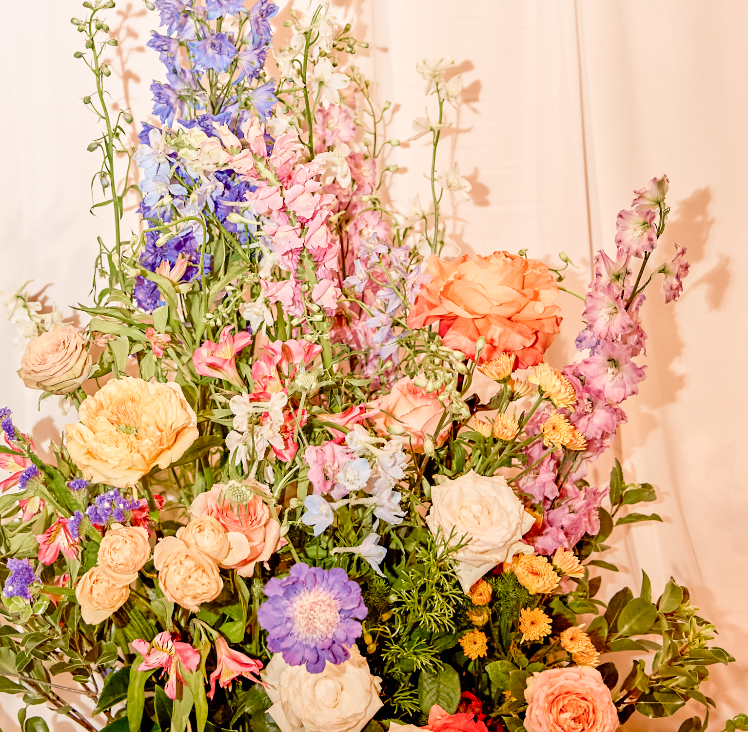 Colorful bouquet of mixed flowers including roses, delphiniums, and other blossoms in shades of pink, purple, orange, and white against a soft pink background.