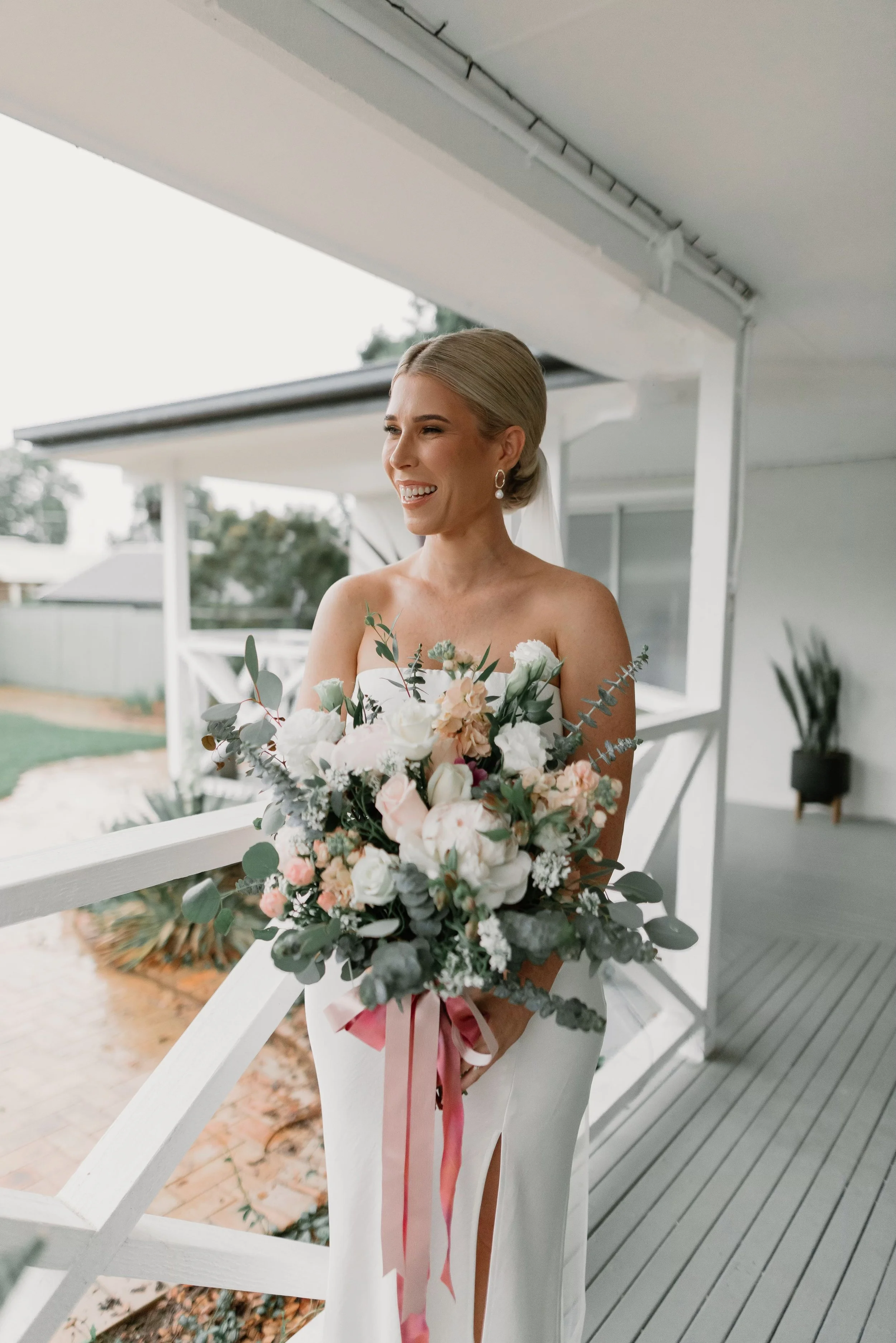 A smiling bride holding a bouquet of flowers, standing on a porch with a white railing and house in the background.