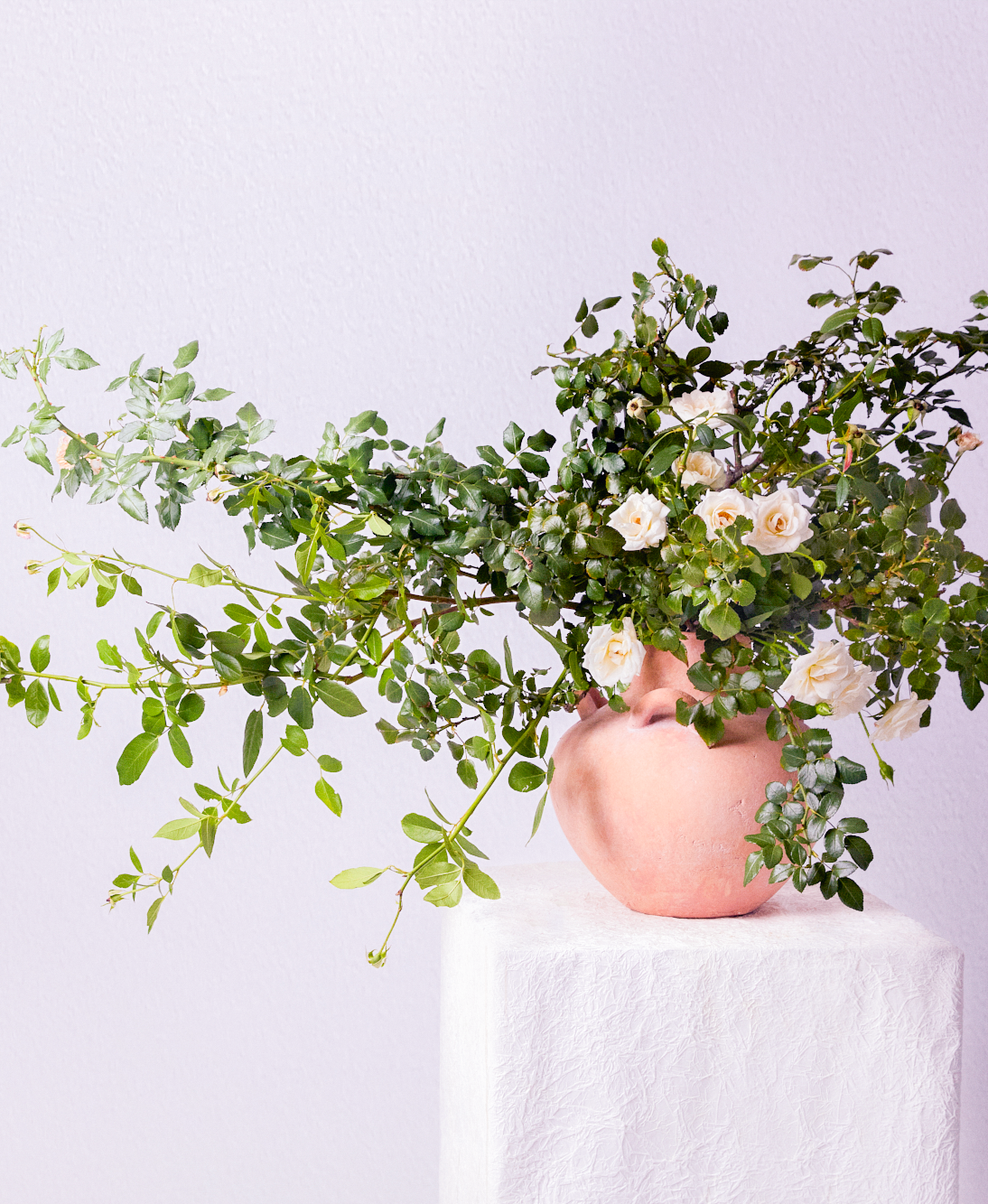 A pink vase filled with lush green foliage and white flowers on a white textured pedestal against a light pink wall.