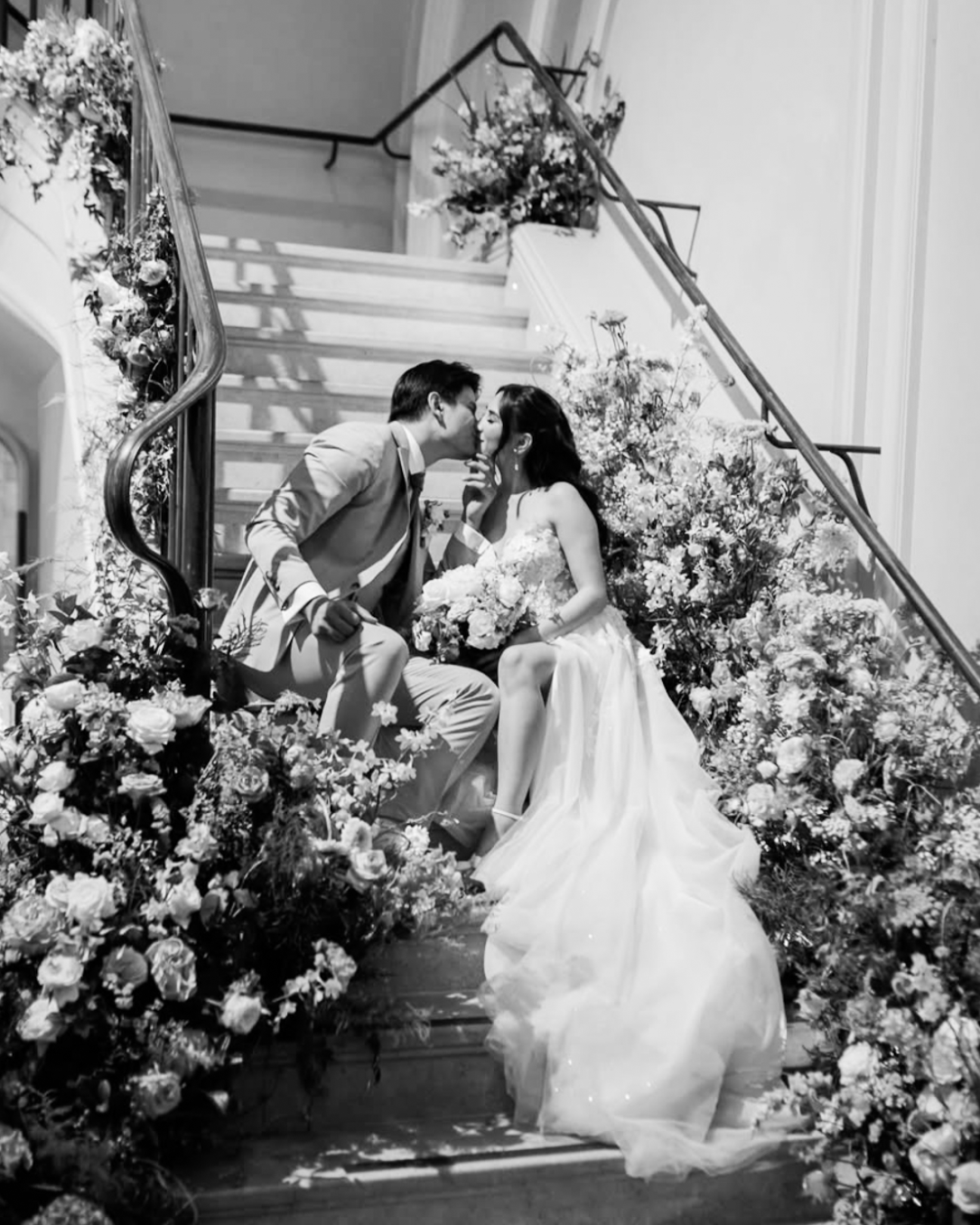 A black and white photo of a wedding scene with a bride in a wedding dress and a groom in a suit sitting on a staircase decorated with flowers. They are leaning in to kiss each other, and the bride is holding a bouquet of flowers.