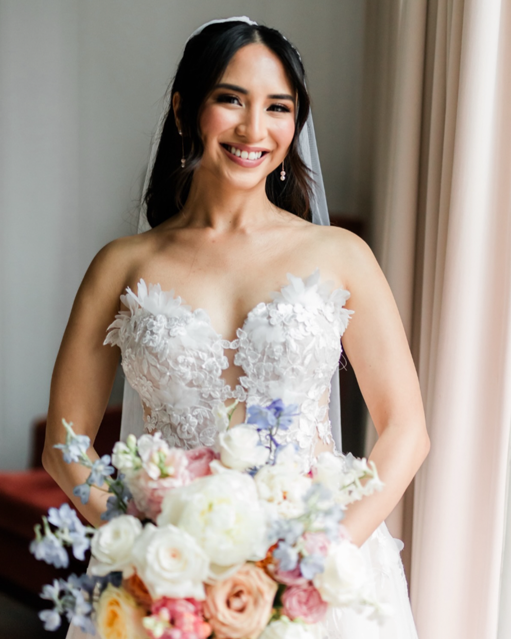 A smiling bride with dark hair in a white wedding dress holding a bouquet of peonies, roses, and blue flowers, standing near a window.