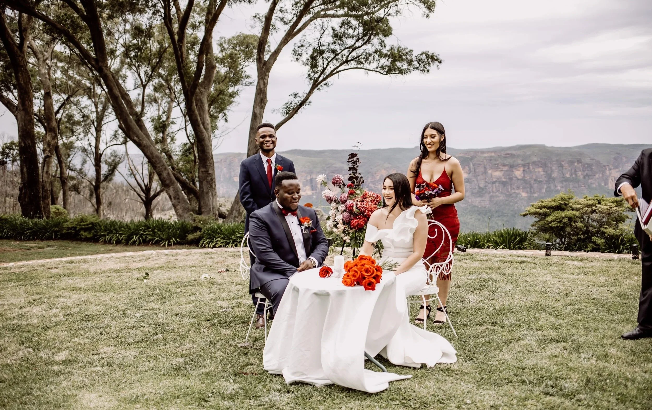 A wedding ceremony outdoors with a bride and groom seated at a table decorated with orange and red flowers. Several guests stand nearby, including two women, one in a red dress and the other in a white dress. Large trees and mountains are in the back