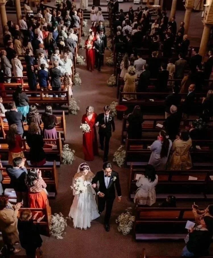 A wedding ceremony inside a church with the bride and groom walking down the aisle, surrounded by guests seated in pews and floral arrangements.