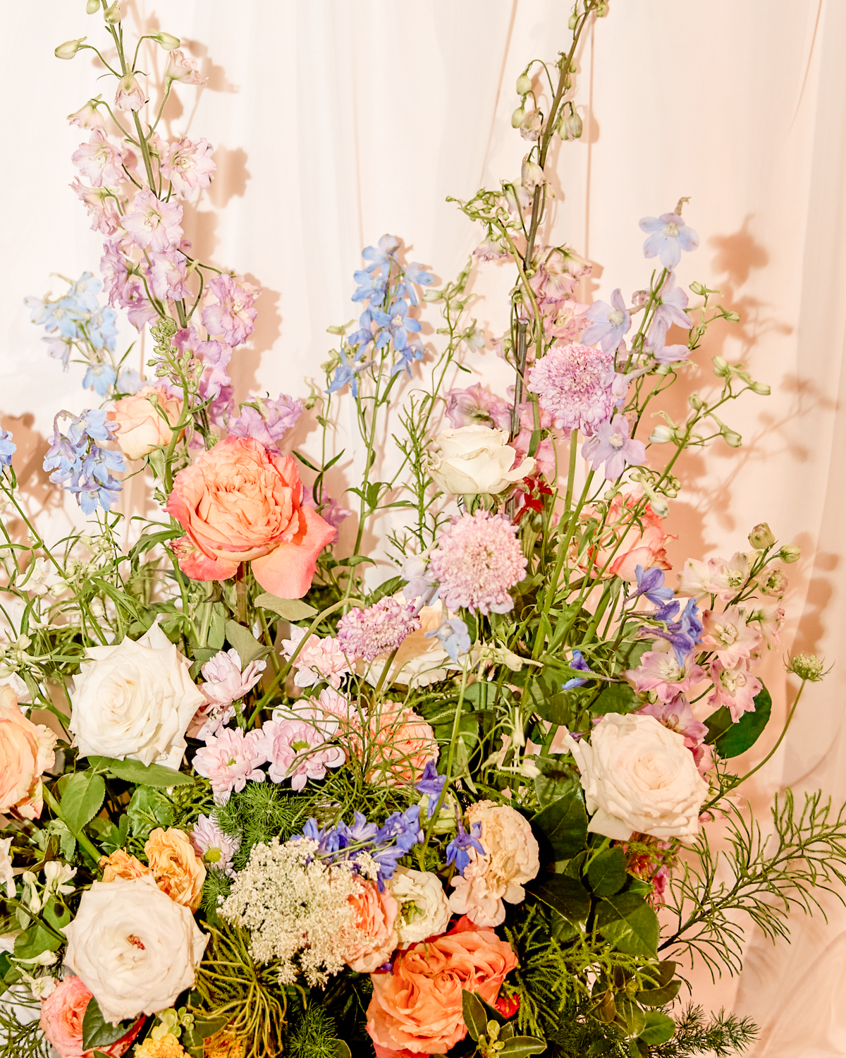 A colorful bouquet of various flowers including roses, delphiniums, and other blooms against a light pink background.
