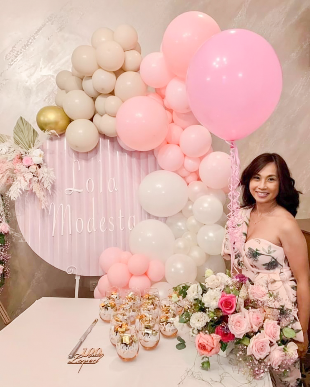 A woman in a strapless pink dress with a floral pattern, smiling, standing near a table with flower arrangements and glass jars filled with candies, at a celebration with pink and white balloons and a decorative backdrop that reads 'Lola Modesta'.
