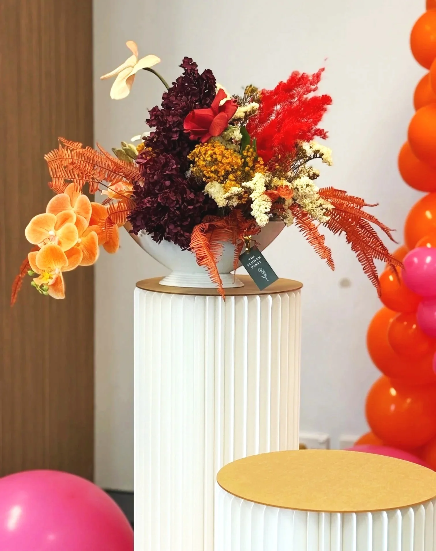 A floral arrangement in a white vase on a white ribbed pedestal, with orange and pink balloons nearby, situated in a room with plain walls and a wooden door.