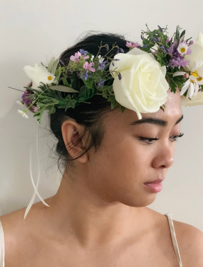 A woman with dark hair wearing a flower crown composed of white roses, daisies, and various small purple and pink flowers, against a plain white background.
