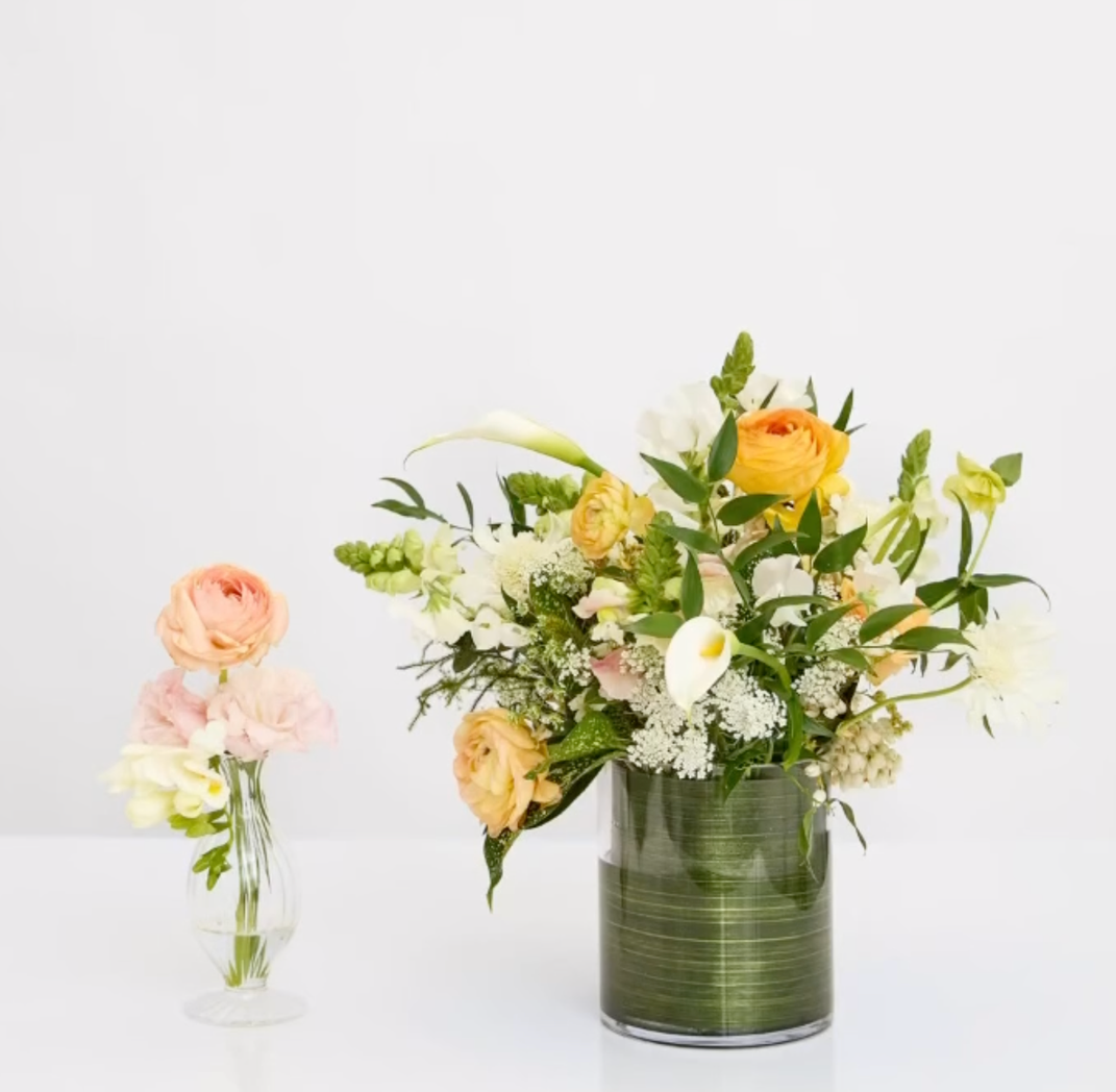 Two vases with flower arrangements: one large green vase with a mix of yellow, white, and green flowers including lilies and roses, and a smaller clear glass vase with pink and white flowers.