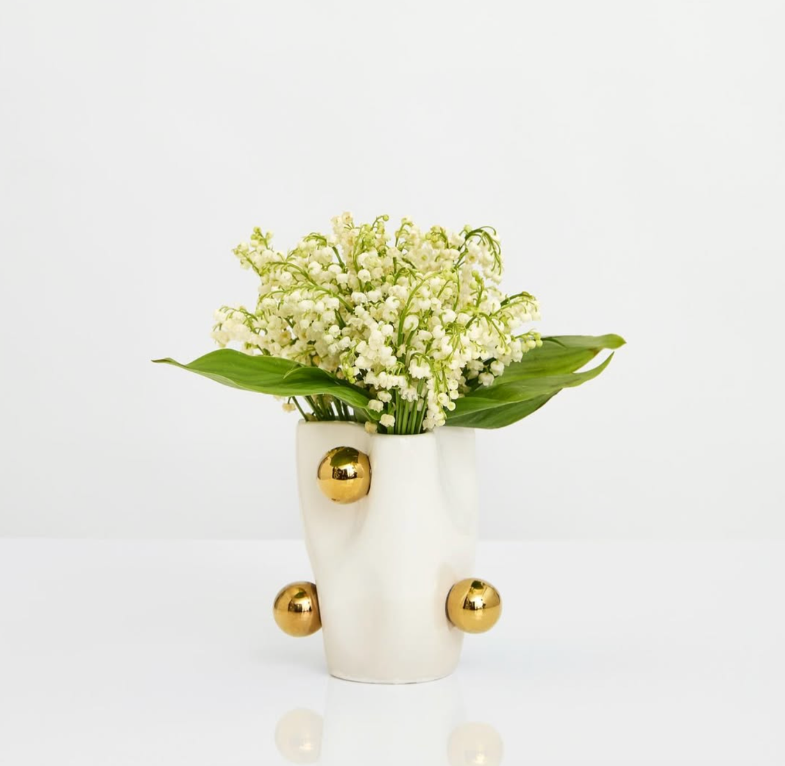 White vase with gold accents holding a bouquet of white lily of the valley flowers and green leaves, placed on a white surface with a plain white background.